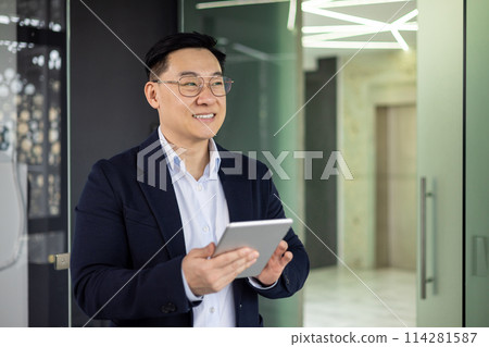 An Asian businessman in a blue blazer smiles as he uses a tablet in a well-lit, contemporary office setting, portraying professionalism and confidence. An Asian businessman in a blue blazer smiles as he uses a tablet in a well-lit, contemporary office setting, portraying professionalism and confidence. 114281587
