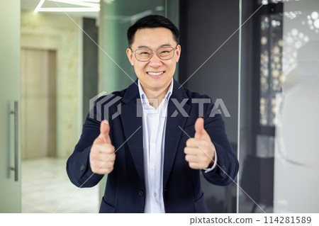 A professional Asian businessman in a navy suit stands in a modern office, smiling and giving a thumbs up, exuding confidence and success. A professional Asian businessman in a navy suit stands in a modern office, smiling and giving a thumbs up, exuding confidence and success. 114281589