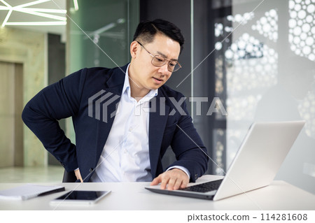 Asian businessman in a modern office feeling a sharp pain in his back while working intensely on a laptop. Focus on health issues related to office work. 114281608