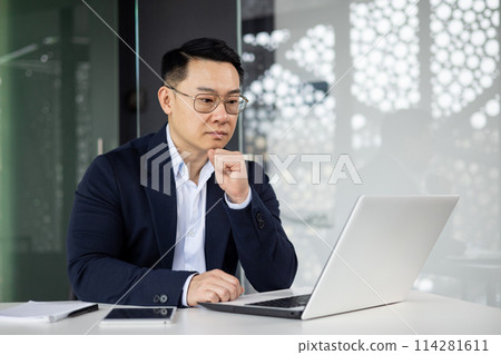 A professional Asian businessman in a blue suit focuses intently on his laptop screen while sitting at a white table in a sleek, contemporary office space. A professional Asian businessman in a blue suit focuses intently on his laptop screen while sitting at a white table in a sleek, contemporary office space. 114281611