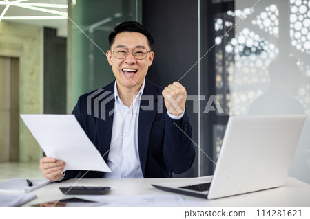 A joyful Asian businessman celebrates a successful deal or achievement at his office desk, showing emotion with a triumphant fist pump. A joyful Asian businessman celebrates a successful deal or achievement at his office desk, showing emotion with a triumphant fist pump. 114281621