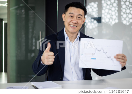 Smiling Asian businessman in a suit giving a thumbs up while holding a financial chart during a video call in a modern office setting. 114281647