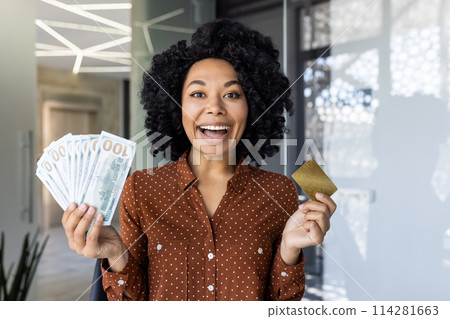 Excited African American woman holding money and a gold credit card, standing in a modern office setting. She displays joy and financial success. 114281663