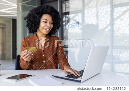 Cheerful young woman with curly hair using a credit card for an online purchase. Sitting at a desk with a laptop in a bright, modern office. 114282112