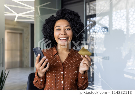 A cheerful African American businesswoman holds a credit card and smartphone in a modern office setting, symbolizing ecommerce and mobile banking. 114282118