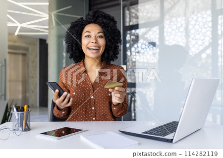 A joyful African-American woman holds a credit card and smartphone, shopping online with a laptop in a modern office setting. 114282119