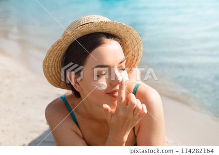 Beautiful Young woman with sun cream on face. Girl holding sunscreen bottle on the beach. Female in hat applying moisturizing lotion on skin.Skin care. Sun protection. Suntan Beautiful Young woman with sun cream on face. Girl holding sunscreen bottle on the beach. Female in hat applying moisturizing lotion on skin.Skin care. Sun protection. Suntan 114282164