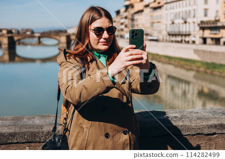 Attractive young female tourist is exploring new city. Redhead girl holding a smartphone on famous Old bridge in Florence. Traveling. Happy optimistic girl walking in city and makes selfie Attractive young female tourist is exploring new city. Redhead girl holding a smartphone on famous Old bridge in Florence. Traveling. Happy optimistic girl walking in city and makes selfie 114282499