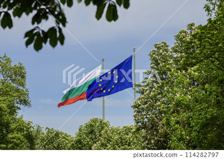Blue flag of European Union and state national Bulgarian flag waving against background of the sky and plant greenery 114282797