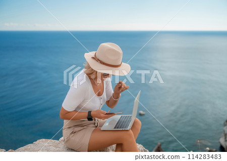 Freelance women sea working on the computer. Good looking middle aged woman typing on a laptop keyboard outdoors with a beautiful sea view. The concept of remote work. 114283483