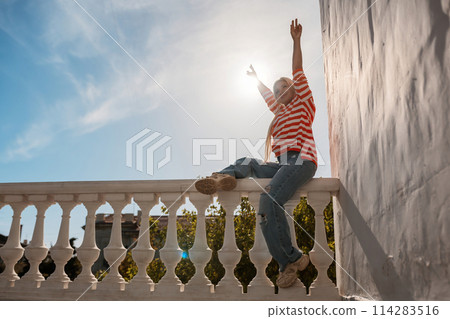A woman is sitting on a railing with her arms raised in the air. The sun is shining brightly, creating a warm and inviting atmosphere. A woman is sitting on a railing with her arms raised in the air. The sun is shining brightly, creating a warm and inviting atmosphere. 114283516