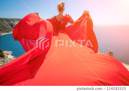 woman sea red dress. Blonde with long hair on a sunny seashore in a red flowing dress, back view, silk fabric waving in the wind. Against the backdrop of the blue sky and mountains on the seashore. woman sea red dress. Blonde with long hair on a sunny seashore in a red flowing dress, back view, silk fabric waving in the wind. Against the backdrop of the blue sky and mountains on the seashore. 114283525