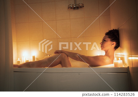 A woman is sitting in a bathtub with candles lit around her. Scene is relaxing and calming. 114283526
