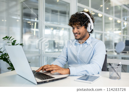 A cheerful young man with curly hair wears headphones while focusing on work using a laptop in a modern office environment, showcasing productivity and concentration. A cheerful young man with curly hair wears headphones while focusing on work using a laptop in a modern office environment, showcasing productivity and concentration. 114283726