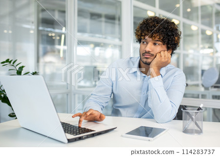 A young professional appears bored and disengaged while working at his laptop in a bright, modern office. He gazes out a window, seemingly lost in thought. A young professional appears bored and disengaged while working at his laptop in a bright, modern office. He gazes out a window, seemingly lost in thought. 114283737