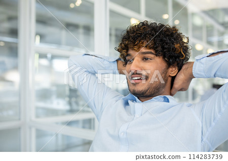 Portrait of young unshaved man in formalwear holding hands behind head while leaning on back of chair. Positive worker looking forward and daydreaming about upcoming vacation on blurred background. 114283739