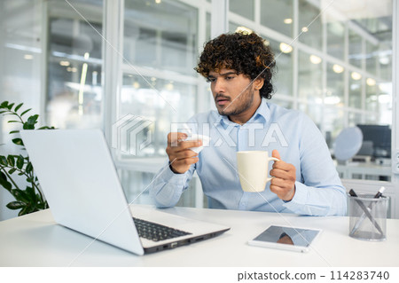 A young businessman with curly hair feeling unwell, holding a tissue and a cup of tea, working on his laptop in a modern office environment. 114283740