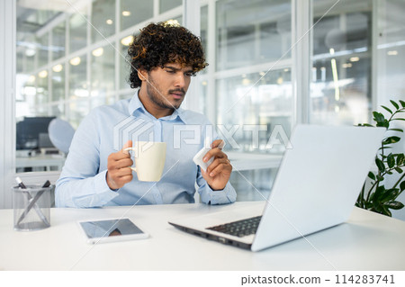 A young adult man in a bright office setting, holding a cup and a thermometer, looking stressed and unwell. He seems to be checking for fever symptoms, depicting a general health concern. 114283741
