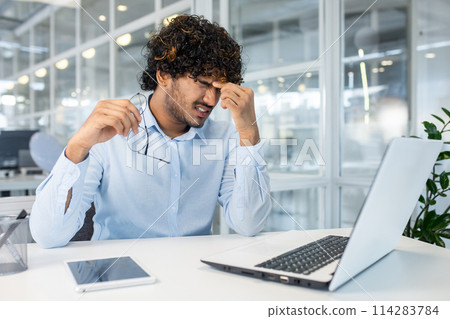 A young man sits at his office desk, visibly frustrated, rubbing his temples in an attempt to alleviate stress or a headache. An open laptop and digital tablet are on the desk. A young man sits at his office desk, visibly frustrated, rubbing his temples in an attempt to alleviate stress or a headache. An open laptop and digital tablet are on the desk. 114283784