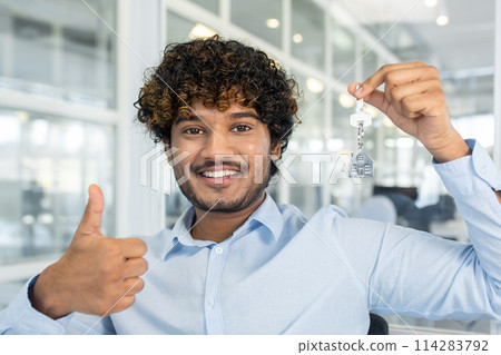 Diverse real estate agent smiling at camera while showing thumb up gesture and holding keys with chain in shape of house. Friendly male recommending buying apartment in new residential complex. 114283792