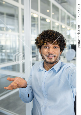 A smiling young man with curly hair makes a video call from a modern office. He's looking at the camera, engaging in a friendly conversation. A smiling young man with curly hair makes a video call from a modern office. He's looking at the camera, engaging in a friendly conversation. 114283793