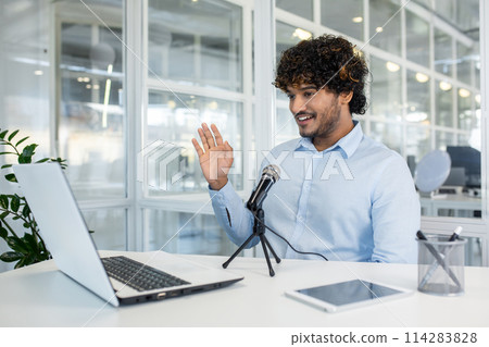 Cheerful young man in a light blue shirt engaging in a virtual business meeting or hosting a webinar from a well-lit office ,space with modern decor. 114283828