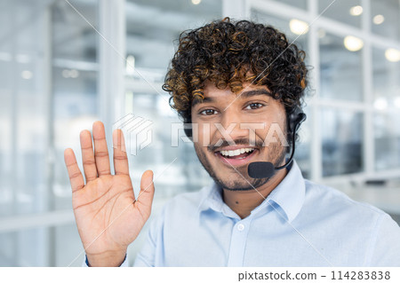 Cheerful young man working in a call center, smiling and waving to the camera, wearing a headset in a bright office environment. Cheerful young man working in a call center, smiling and waving to the camera, wearing a headset in a bright office environment. 114283838