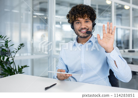 A cheerful call center representative waves at the camera, wearing a headset and holding a digital tablet in a modern office setting. A cheerful call center representative waves at the camera, wearing a headset and holding a digital tablet in a modern office setting. 114283851