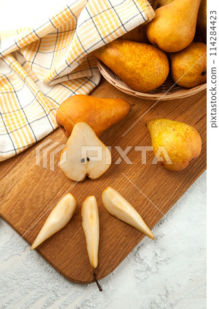 Cutting board with whole and slices pears, wicker basket and yellow kitchen towel on white wooden background.. 114284423