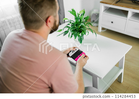 Cheerful young man seated on a sofa playing video games at home. Gaming addiction and entertainment Cheerful young man seated on a sofa playing video games at home. Gaming addiction and entertainment 114284453
