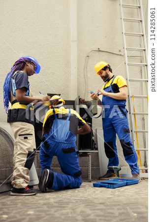 Diverse crew hired by home owner to service faulty air conditioner. Coworkers inspecting defective blower fan causing frozen evaporator coil, resulting in reduced cooling performance 114285061