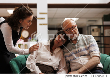 In hotel reception, female concierge waking up elderly couple on comfy sofa for their room reservation. Resort employee awaking sleeping senior travelers in lounge area for their check-in procedure. 114285091