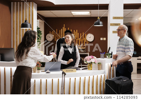 In hotel reception, elderly male and female travelers at registration counter awaiting reservation procedure. Retired senior woman giving personal documents to concierge for check-in. In hotel reception, elderly male and female travelers at registration counter awaiting reservation procedure. Retired senior woman giving personal documents to concierge for check-in. 114285093