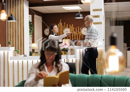 Senior caucasian man standing at hotel lobby counter and checks in with passport. Female concierge at registration desk returning personal document of elderly male traveler after verfiying identity. 114285095