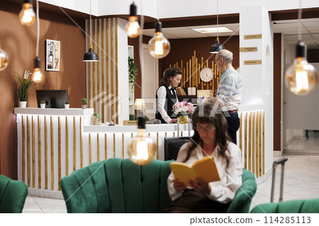 Elderly male guest arrives at front desk of hotel lobby, greeted by friendly female receptionist. Senior woman in cozy lounge area sitting on comfy sofa and reading a book while waiting for check-in. 114285113