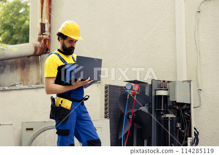 Trained mechanic commissioned for external hvac system routine maintenance, looking for damaged electric components. Meticulous electrician doing condenser safety check, writing report on laptop Trained mechanic commissioned for external hvac system routine maintenance, looking for damaged electric components. Meticulous electrician doing condenser safety check, writing report on laptop 114285119