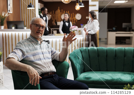 Portrait shot of old man seated on cozy sofa in exclusive hotel reception, waving at camera. Comfortable lobby offers relaxing lounge area for elderly male traveler waiting for check-in procedure. 114285129