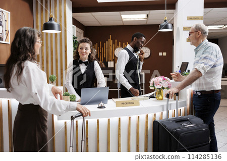 Two elderly travelers come at the hotel reception desk to check in. Multiethnic receptionists assist retired senior man and woman with booking and paperwork, therefore creating a pleasant environment. 114285136