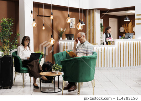 Retired elderly woman sitting on a sofa in the hotel lobby, and using her laptop. Senior male client reading a book in lounge area, while older lady is browsing on her wireless computer. 114285150