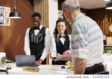 Retired senior male traveler at front desk seamlessly checking in, pays via smartphone in hotel lobby. Friendly concierge at registration counter helps old man with nfc payment using mobile device. Retired senior male traveler at front desk seamlessly checking in, pays via smartphone in hotel lobby. Friendly concierge at registration counter helps old man with nfc payment using mobile device. 114285153