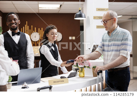 Elderly male tourist at counter using credit card to pay for reservation in exclusive hotel lobby. Multiethnic employees assist caucasian guests with payment and document verification for check-in. Elderly male tourist at counter using credit card to pay for reservation in exclusive hotel lobby. Multiethnic employees assist caucasian guests with payment and document verification for check-in. 114285179