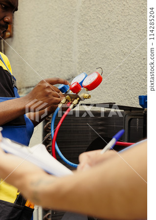 Professional workers assigned to do air conditioner check, refilling freon. Expert repairmen team using ac gauges vacuum pump to skilfully measure the pressure in HVAC system 114285204