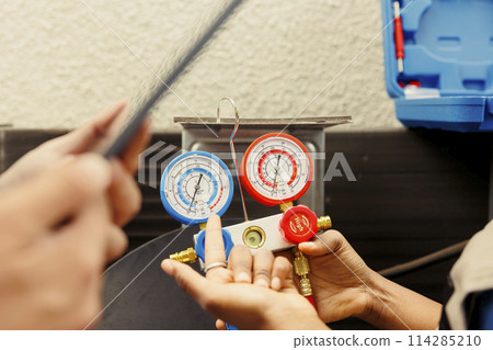 Close up of seasoned wireman holding pressure measurement device used for checking air conditioner refrigerant tank in need of repairing. Proficient expert using manometers to check freon levels Close up of seasoned wireman holding pressure measurement device used for checking air conditioner refrigerant tank in need of repairing. Proficient expert using manometers to check freon levels 114285210