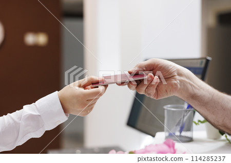 Close-up shot of caucasian pair of hands grasping passport for check-in verification procedure. In hotel reception, senior man handing his personal document to concierge for reservation. Close-up shot of caucasian pair of hands grasping passport for check-in verification procedure. In hotel reception, senior man handing his personal document to concierge for reservation. 114285237