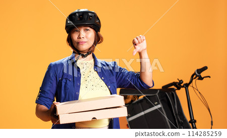 Close up shot of part time courier holding takeaway pizza boxes, showing thumbs down sign. Food delivery cyclist doing disapproving finger shaking gesture, saying no, isolated over studio background 114285259