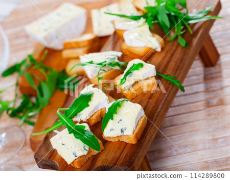 On table there is cutting board with blue cheese and hardtack decorated with arugula On table there is cutting board with blue cheese and hardtack decorated with arugula 114289800