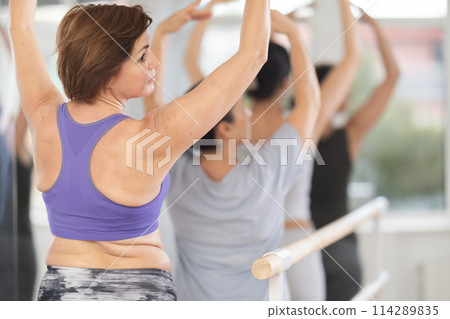 Ordinary women, not professionals, stand in fifth position near ballet barre during group training in dance studio Ordinary women, not professionals, stand in fifth position near ballet barre during group training in dance studio 114289835