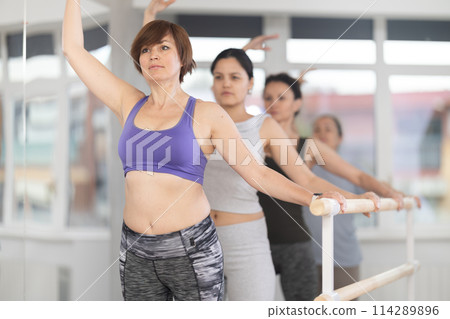 Female amateurs stand in third position near ballet barre during group training in dance studio Female amateurs stand in third position near ballet barre during group training in dance studio 114289896