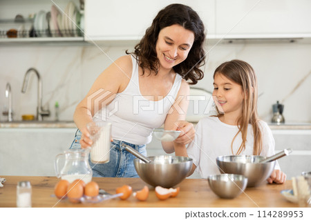 Happy mother and her daughter adding flour in metal bowl and whisking ingredients in the kitchen 114289953