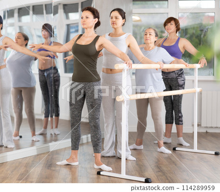 Women of different ages exercise near the ballet barre at group training session in batman batyu position Women of different ages exercise near the ballet barre at group training session in batman batyu position 114289959